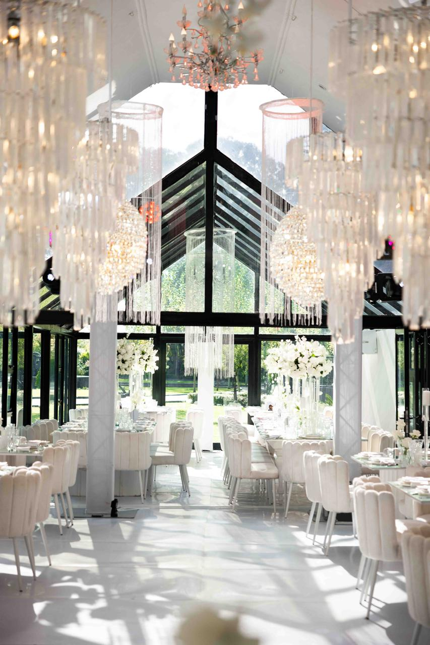Glass-walled reception venue with chandeliers and white seating